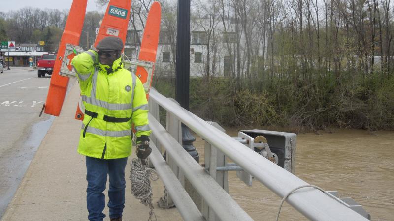 USGS employee using water monitoring equipment over a bridge in an urban area