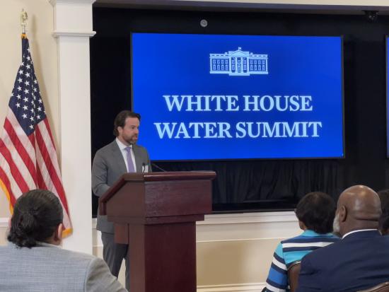 Man standing behind podium next to American Flag and Sign that says White House Water Summit