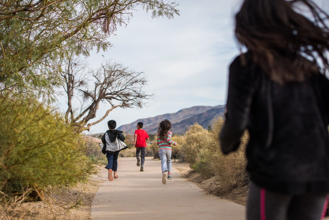 Four young children run along a walkway with desert plants growing on either side.