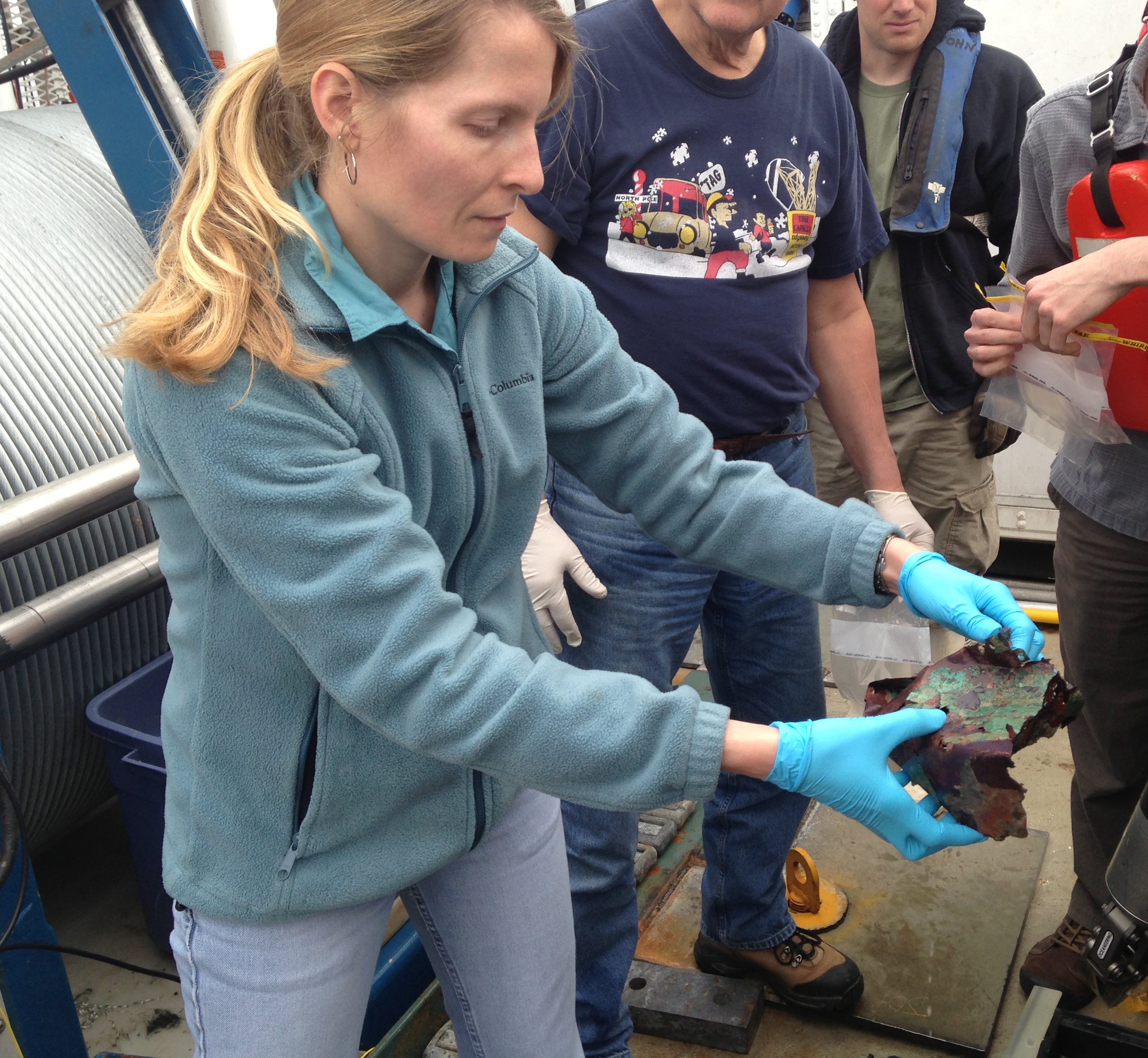 Melanie holds a metal sample in gloved hands with a group of people in the background.