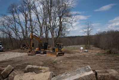 Construction vehicles clear an open, rural site after a demolition, with trees behind.