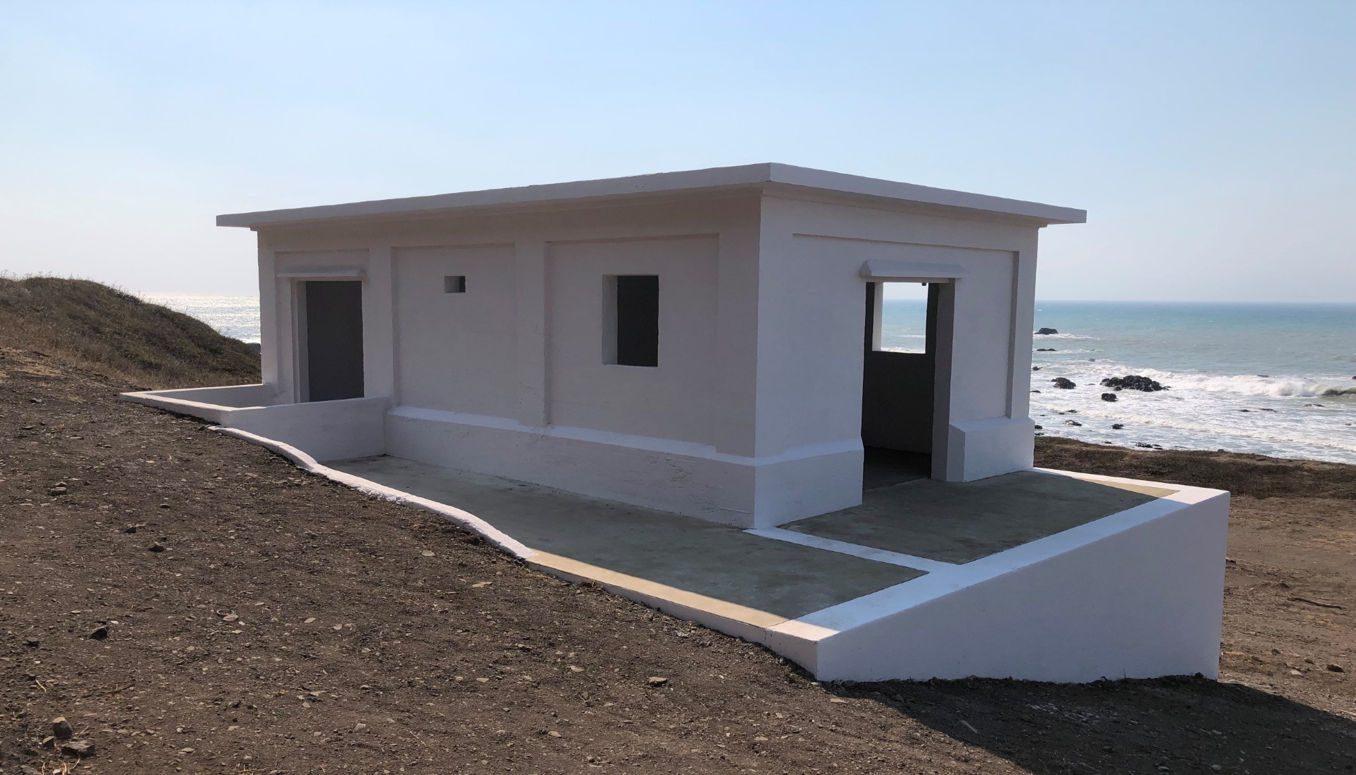 A small white building sits on a brown sandy cliff with the blue ocean in the background