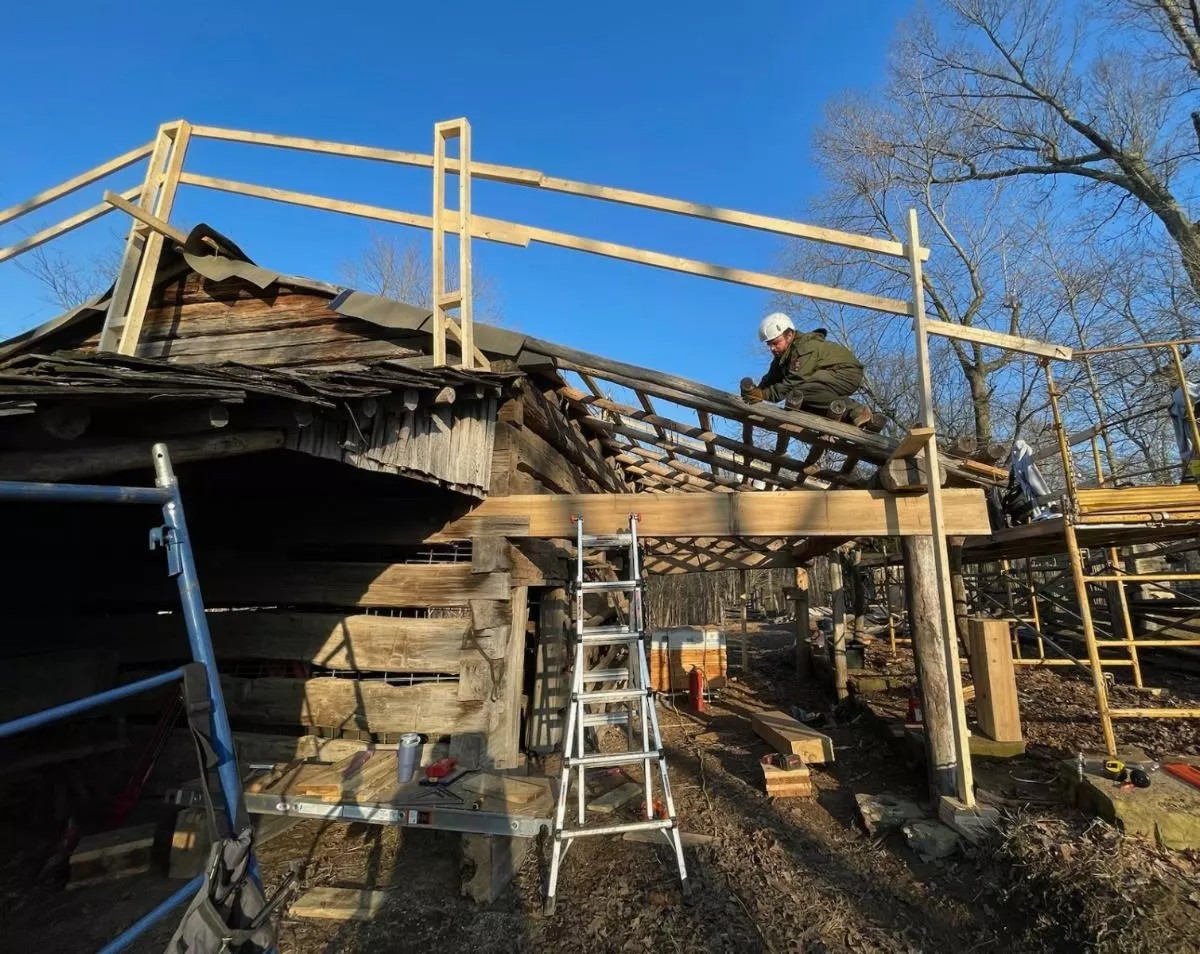 Worker in safety gear putting new wooden roof on a log barn with a bright blue sky in the background