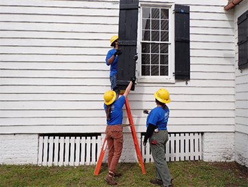 Three workers in yellow construction hats replace a black window shutter on a white building