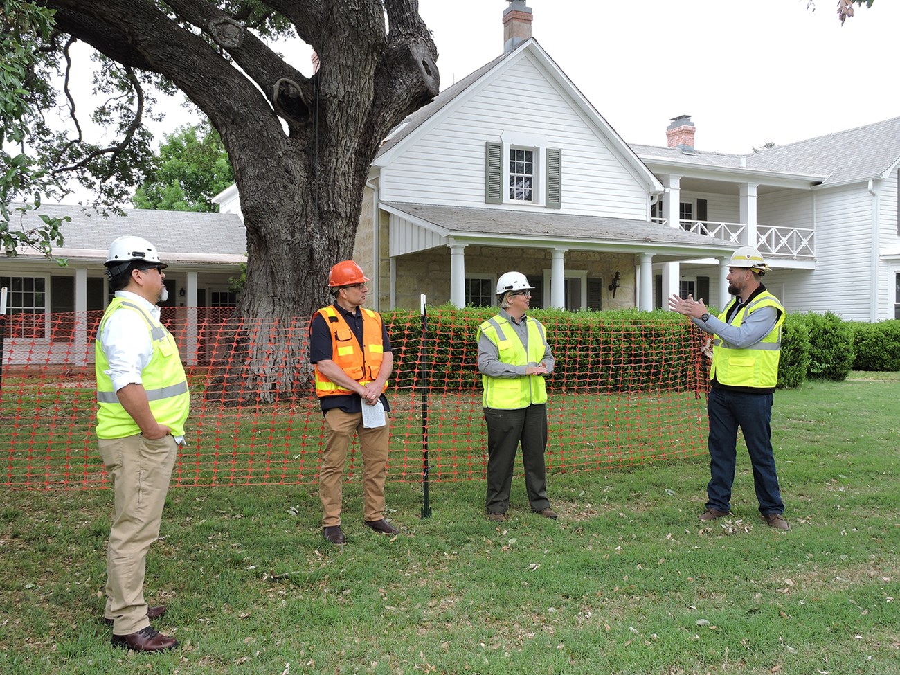 People in construction vests stand in front of a white house surrounded by orange fencing