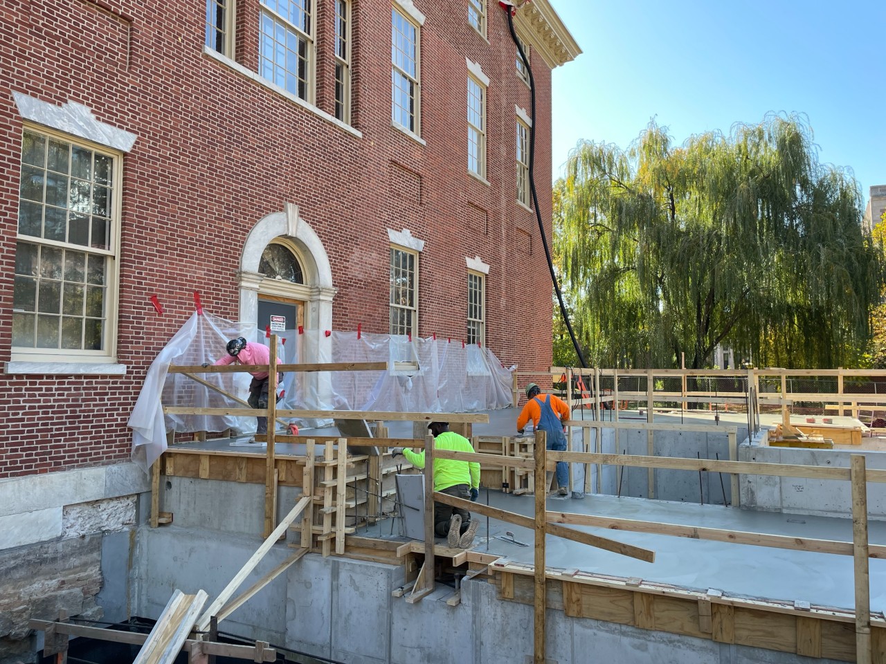 Construction workers stand on concrete platform with wooden railings attached to the exterior of brick building.