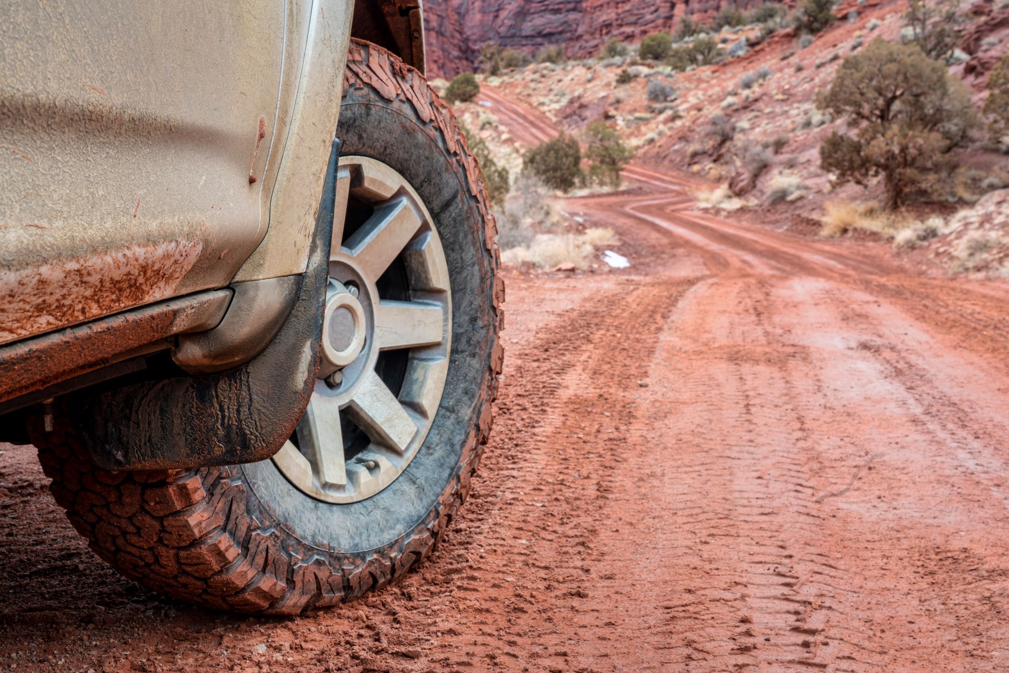 A SUV is on a dirt road. You can see the front tire of the vehicle and the red dirt road in front of it. 