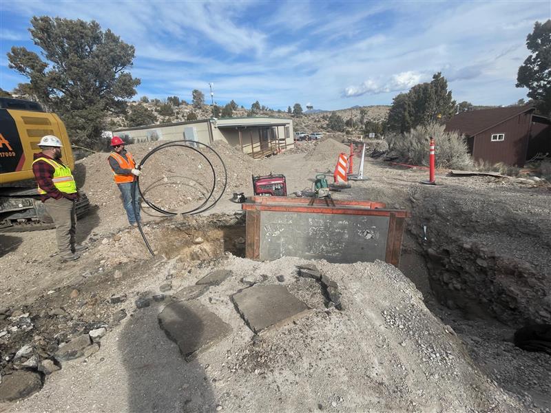  Two men in hard hats stand next to a large dirt hole with water system components inside.