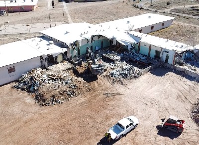 An excavator demolishes an education building on a dirt plot.