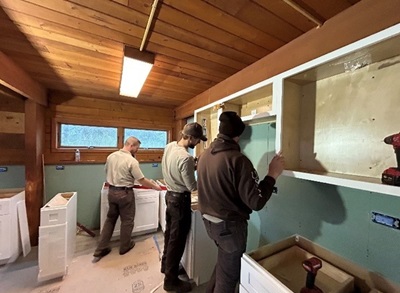 Three MAT members hang cabinets in a room with green walls and board ceiling.