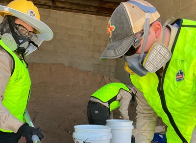 Three people wearing masks and yellow construction vests sift through sand in a metal tray.