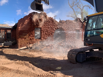 Yellow construction vehicle demolishes red brick building in construction area.