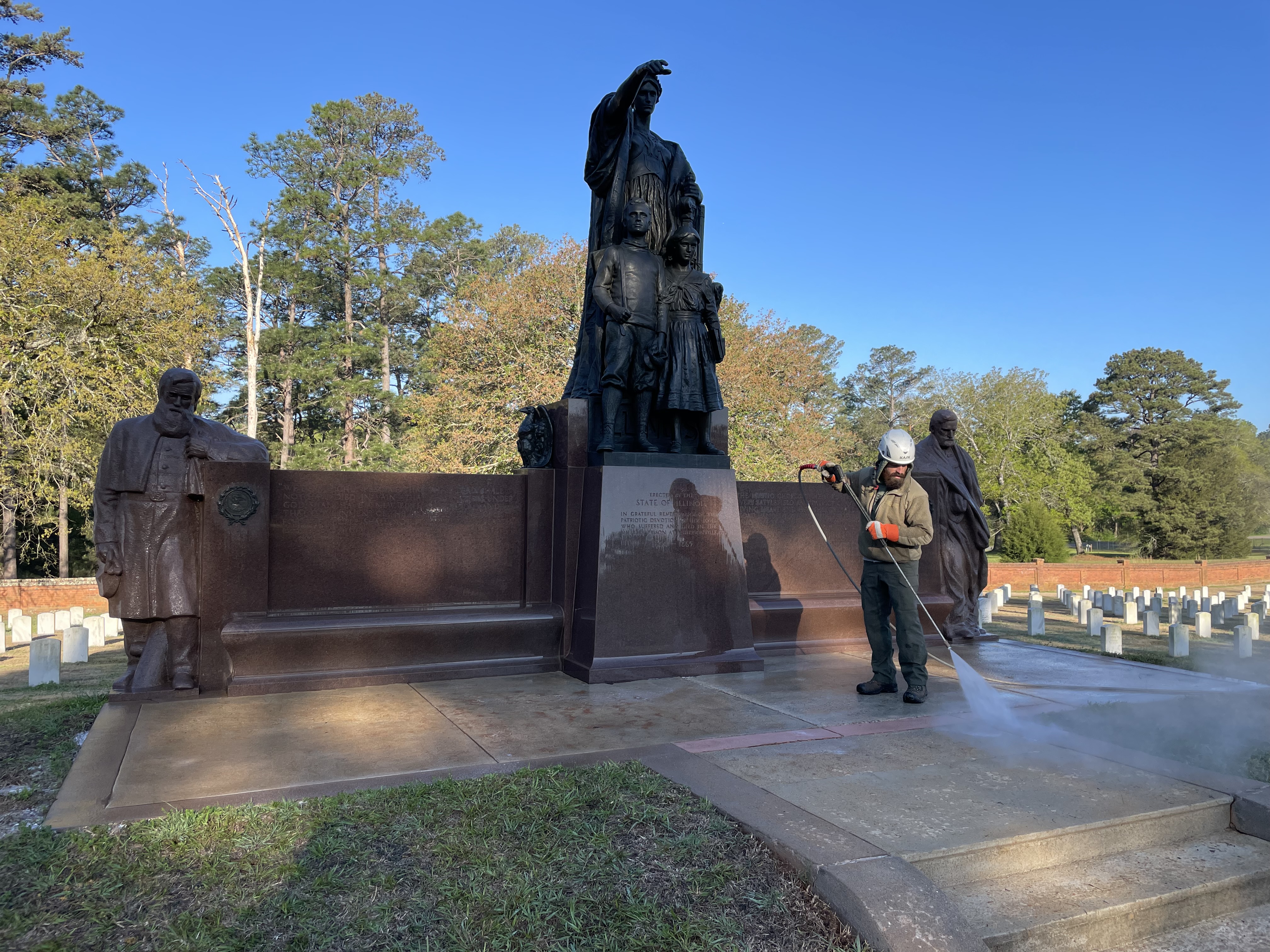 Man in white construction hat hoses the concrete patio in front of a statue.