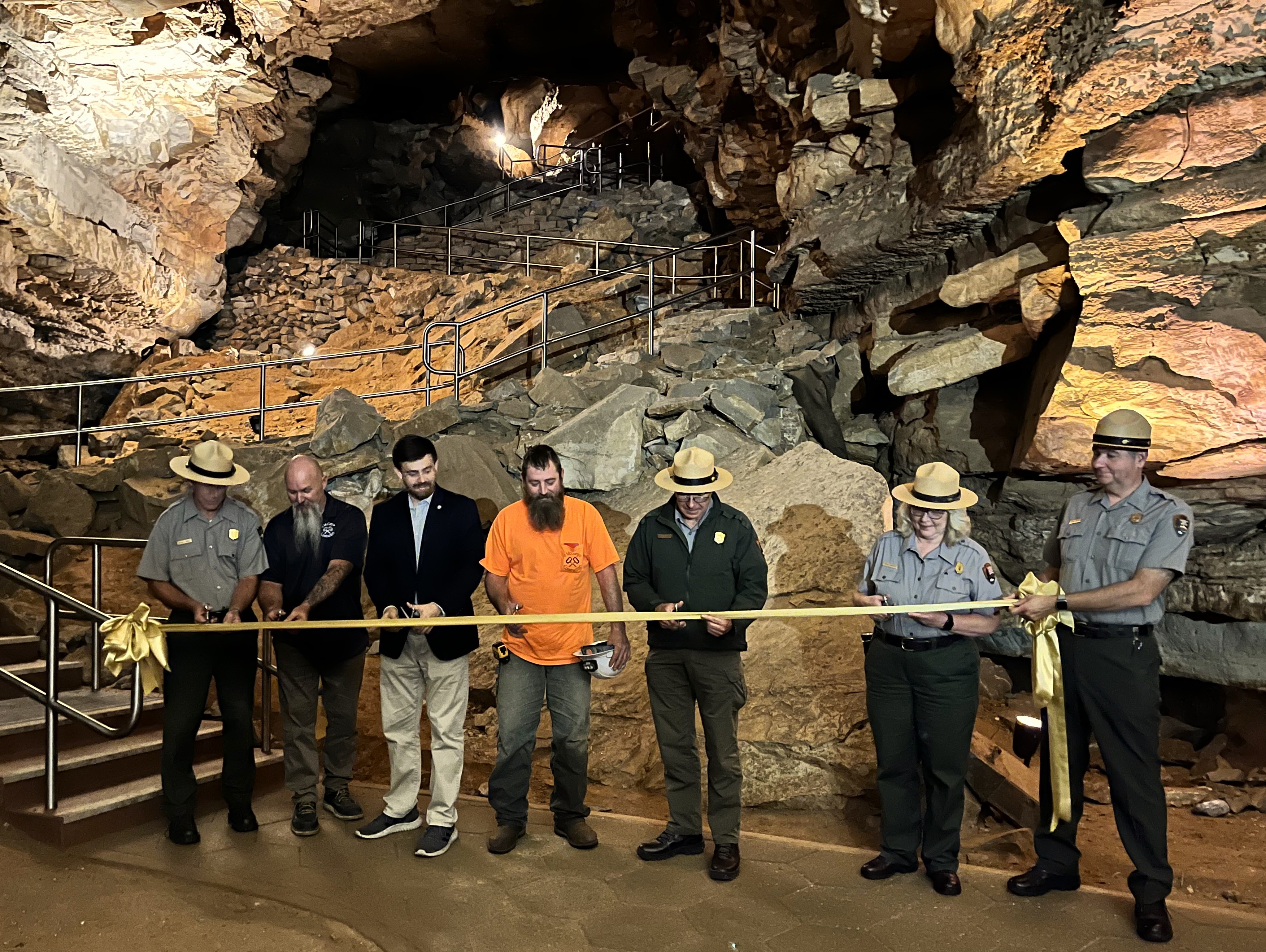  Seven people stand behind gold ribbon on newly rehabilitated cave trail.