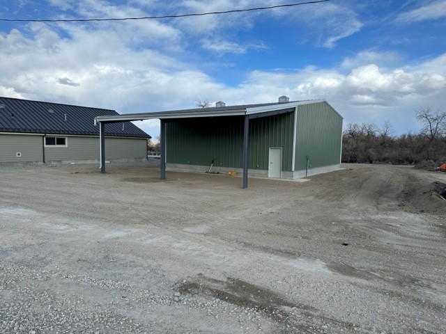 New, green metal utility building with an open covered bay, set on a gravel lot beside another green facility building.