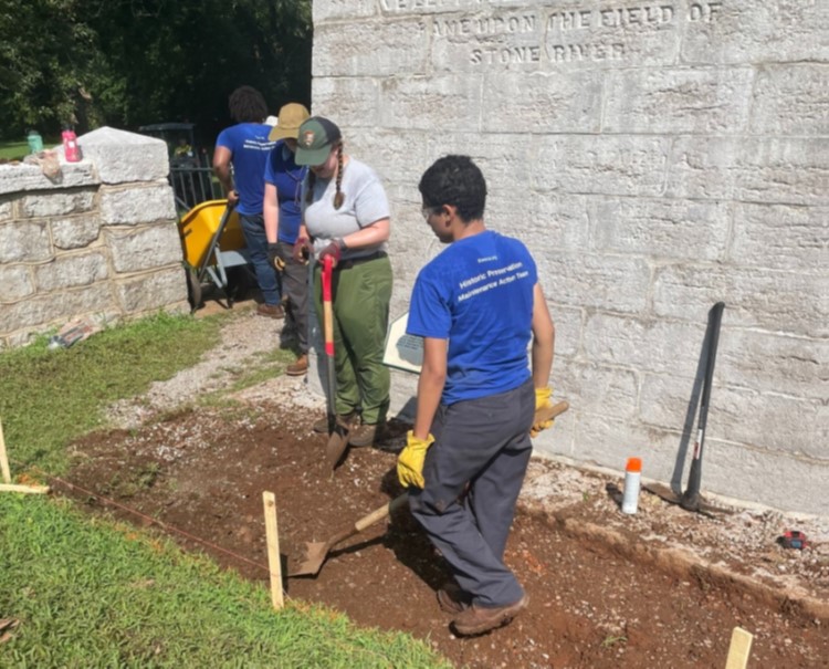 Student volunteers use shovels and tools to place and level soil along the base of a stone wall during site restoration work.