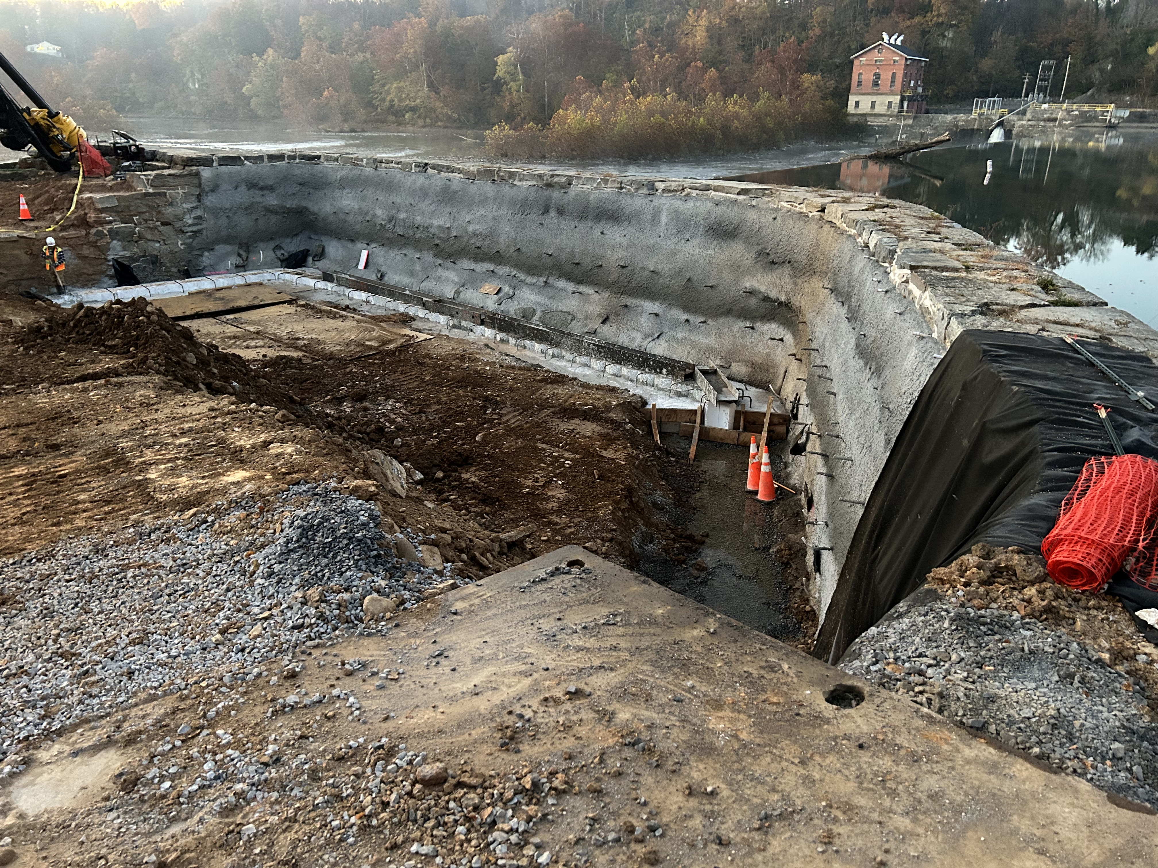  Excavation and concrete repair work along a curved stone dam, with a canal visible in the background.