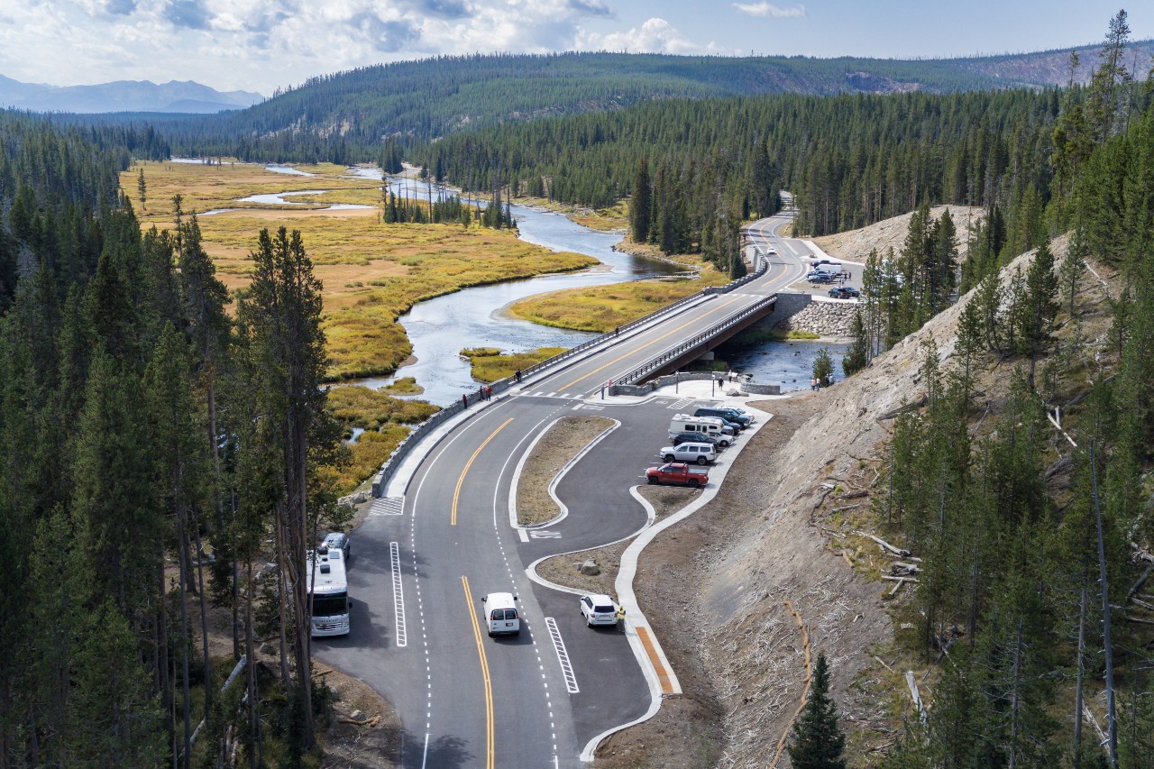 Newly constructed bridge crosses a winding river surrounded by mountains and tall trees.