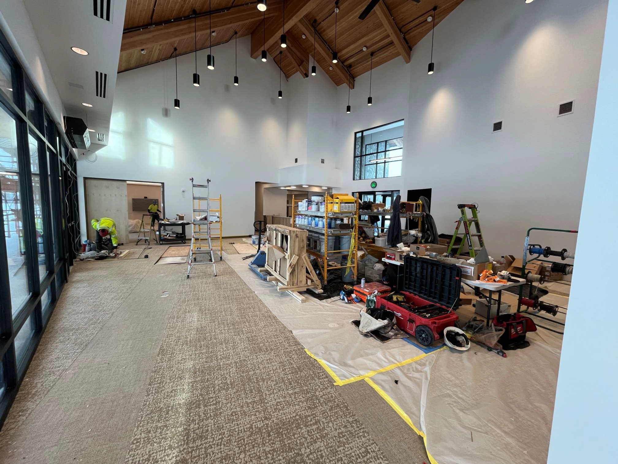  Interior view of a large, high-ceilinged multipurpose space under construction, featuring exposed wooden beams, hanging light fixtures, and large windows.