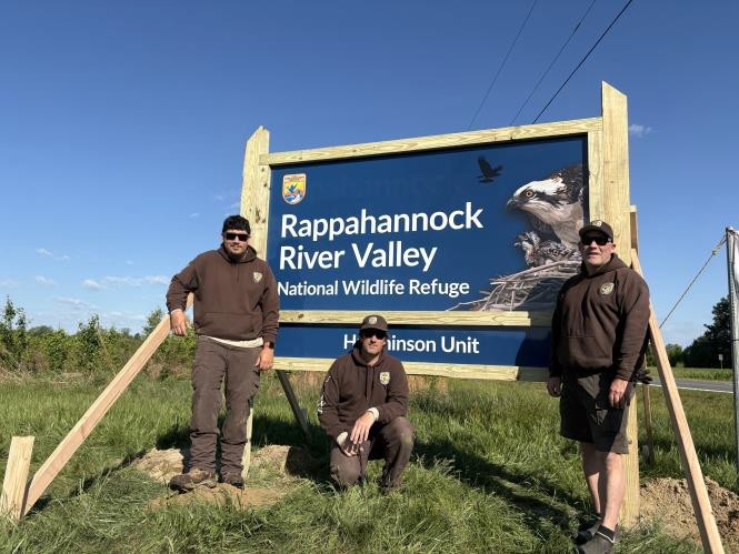 Maintenance Action Team workers stand beside a newly installed “Rappahannock River Valley National Wildlife Refuge” sign in a grassy roadside area.