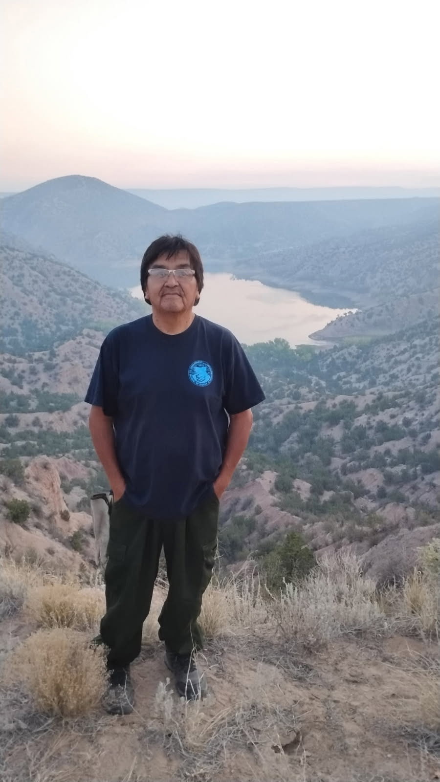 Randy Lupe stands on a hill overlooking a small lake surrounding by hills and shrubland under a hazy sky.