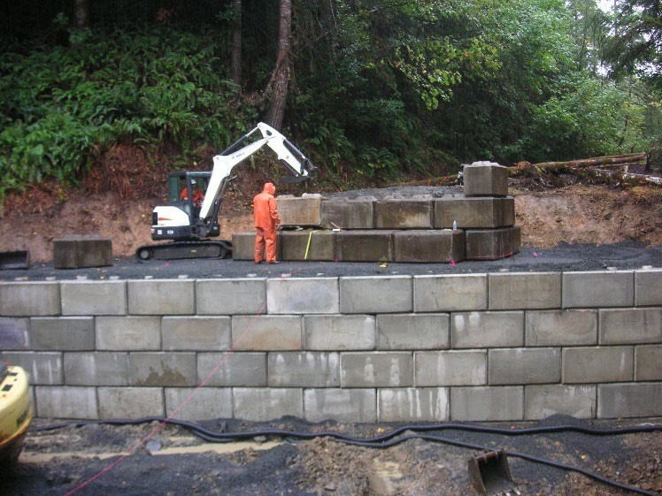 Construction crew members with excavator placing large concrete blocks for a retaining wall.