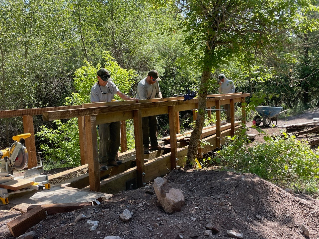 Three maintenance workers build a wooden pedestrian bridge along a dirt path.