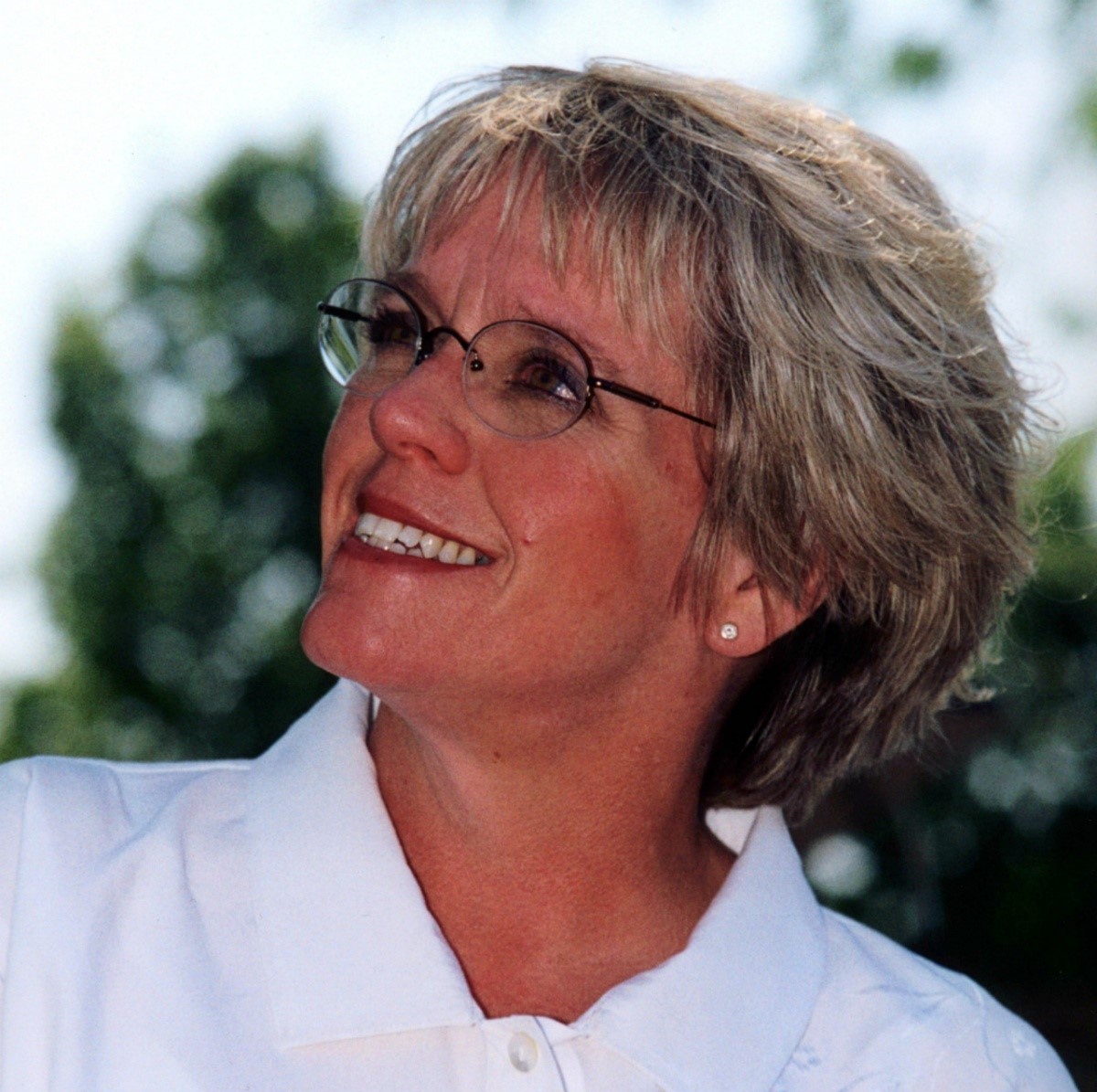 Kathleen Clark, a blonde woman wearing a white shirt, stands outside and looks up at the sky.