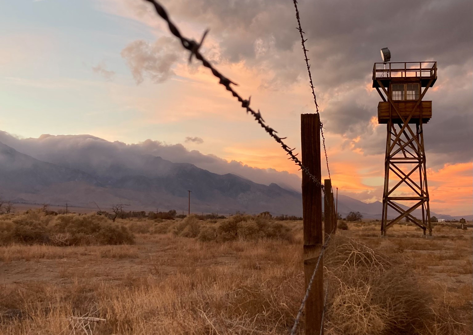 Sunset over guard tower and fence surrounded by mountainous landscape.