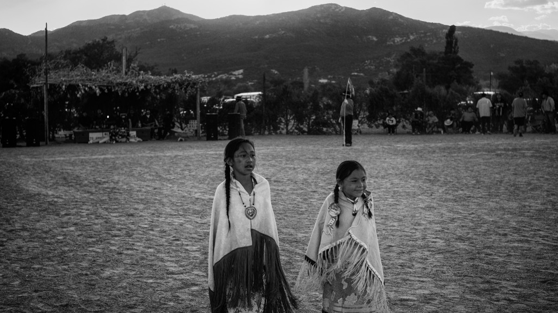 Two Native American girls stand in a field with mountains in the background.