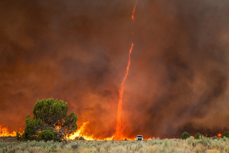 Truck heads out across a field, away from a fire whirl and smoke.