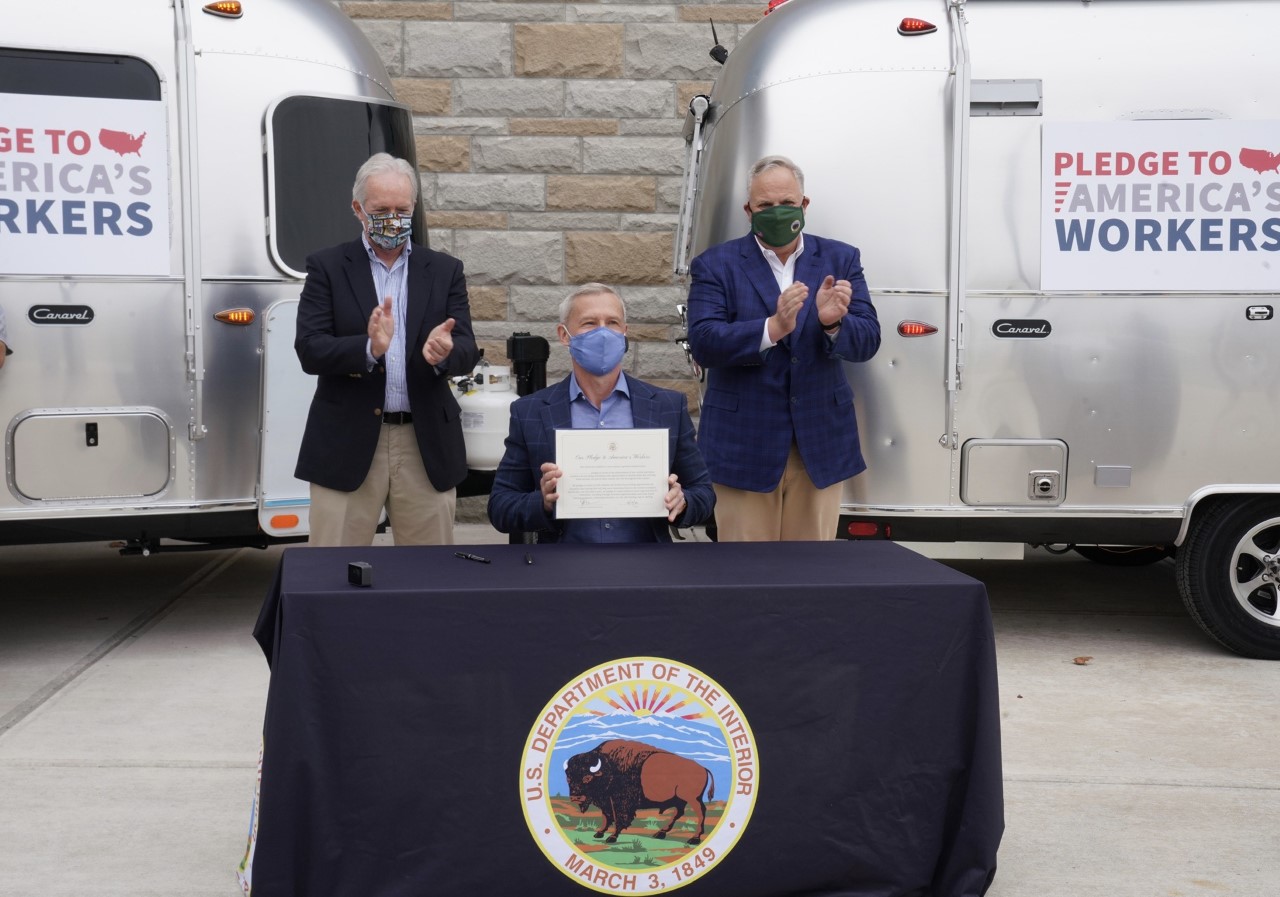 With two metal RVs in the background, Secretary Bernhardt looks on as a man sits at a table and holds up a piece of paper.