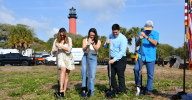  Four people use shovels to break ground on grass with a red lighthouse rising above the trees in the background.