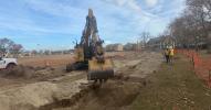 An excavator fills trench with dirt surrounded by temporary orange fencing.