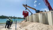 Three men in hard hats stand next to concrete walls blocking a large sand pile and construction vehicles.