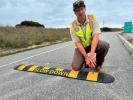 Man in yellow construction vest drills yellow and black speed bump on paved road.