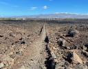 A worn trail is cut through a rough gray and red lava flow, extending in a straight line to the horizon. A low mountain is in the background with blue sky above.