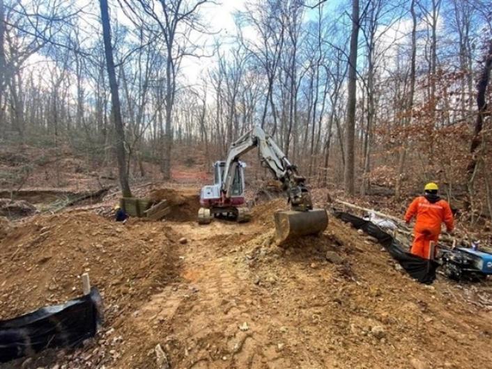 Excavator pulls up dirt in wooded area surrounded by temporary fencing. 
