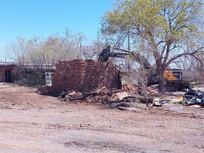 Active demolition of a red brick building with a bulldozer positioned behind a large tree.