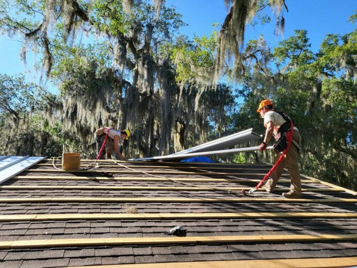 Two women in hard hats stand on the roof of a building and lift metal beams.