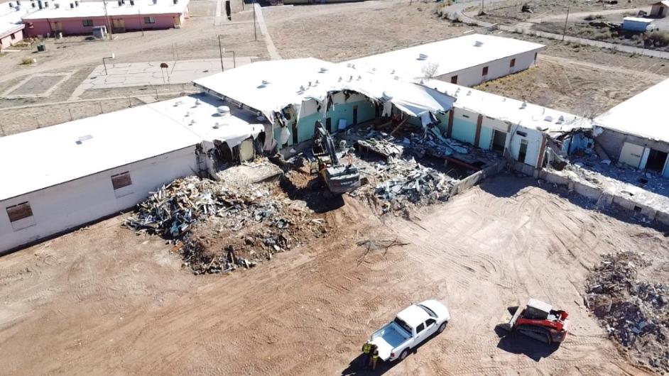 An excavator demolishes an education building on a dirt plot.