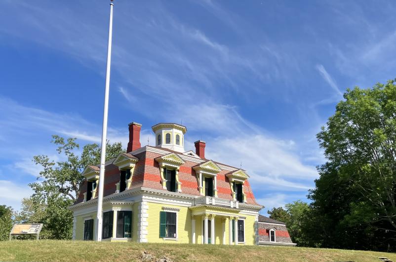 Two story house with yellow siding and red-striped roof on grass lawn.