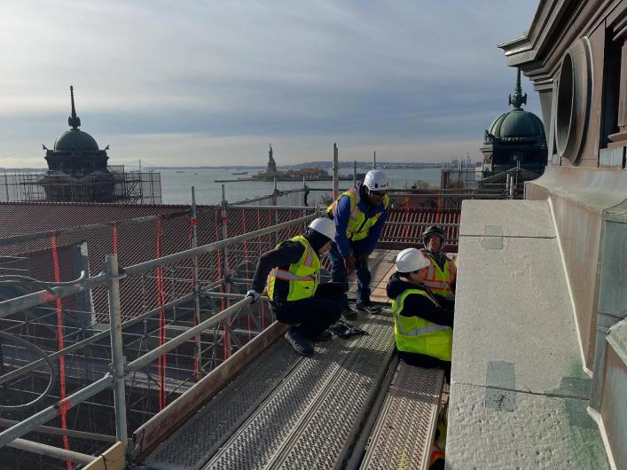 Construction workers repair concrete building exterior with body of water and Statue of Liberty in the background