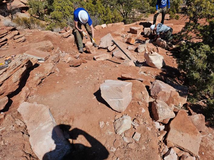 Men in construction hats build delineation walls on canyon trail surrounded by bushes.