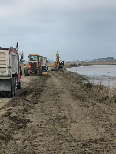 Construction vehicles sit alongside levee leading to a body of water.