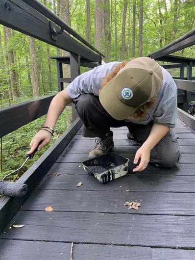 Conservation corps member paints a dark brown, wooden boardwalk