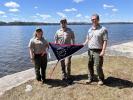 3 Park Rangers holding flag in front of lake front. 