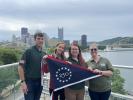 Four people holding commemorative flag in front of Pittsburg skyline. 