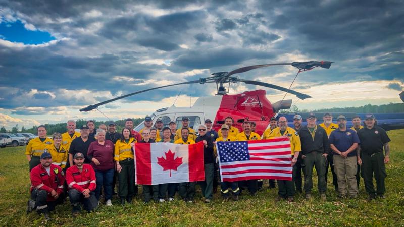 A group of people in different yellow, red, black, and blue uniforms pose for a photo in the middle of a grassy field. Those in the center hold up an American flag and a Canadian flag. A helicopter is parked immediately behind them. A row of parked cars is just visible at the edge of the field to the left. Int eh background, dense trees line the far edge of the field. The sky is a moody patchwork of dark clouds, blue sky, and light clouds illuminated in gold by the setting sun.