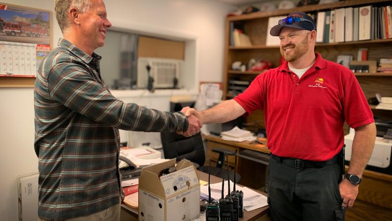 Two men in casual attire shake hands, smiling at each other. Under their clasped hands is a row of radios and a box with more equipment sitting on the corner of a desk. They are standing in a small office. The desk is scattered with papers. The shelves along the back wall hold binders and supplies. On the wall to the left, a calendar displays a photo of a red fire truck.